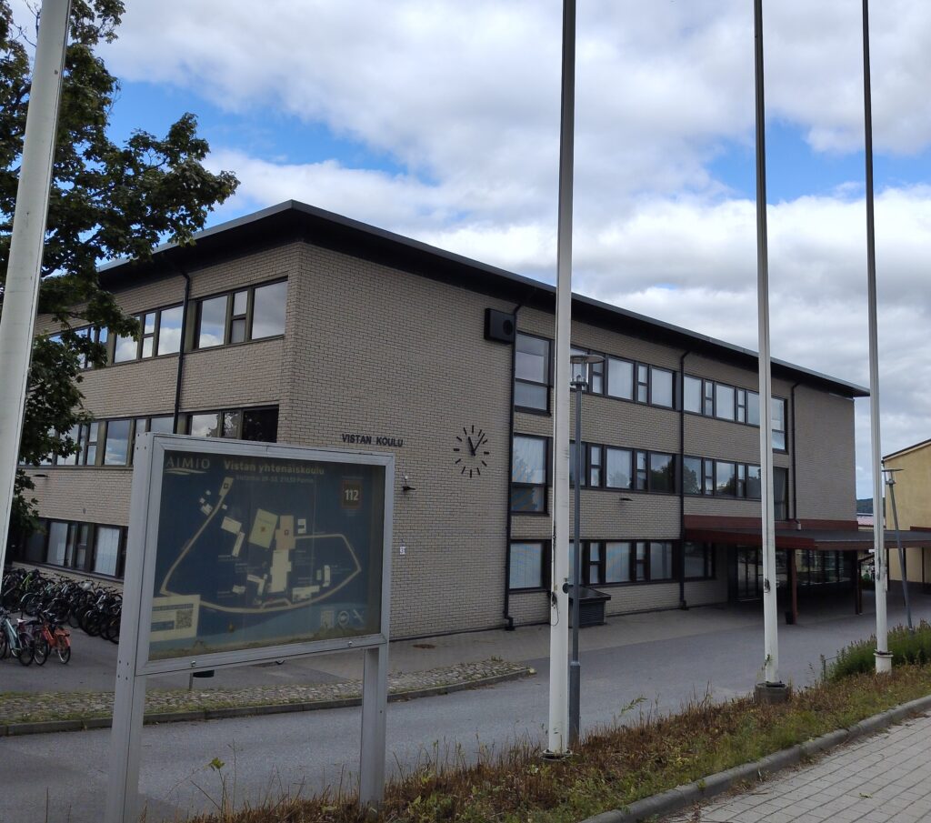 Harmaan, kolmikerroksisen rakennuksen edessä on opastetaulu ja lipputankoja.