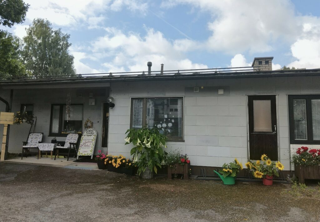 Hyvän Olon Pysäkin rakennus kuvattuna pääsisäänkäynnin puolelta. Kuvassa näkyy myös erilaisia kukkia ja tuoleja.