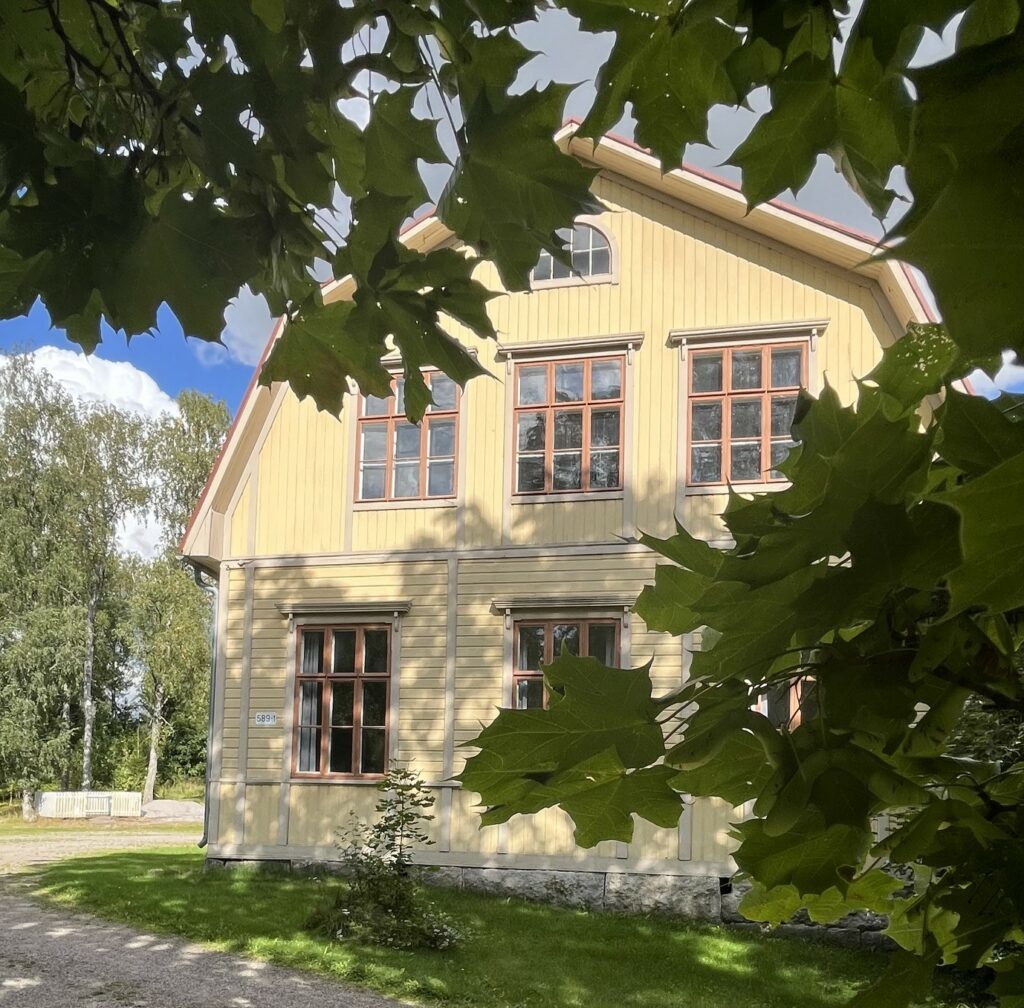 Puolitoistakerroksinen puurakennus, jonka seinässä paljon ruutuikkunoita. Etualalla näkyy vihreitä puiden lehtiä puiden oksilla.