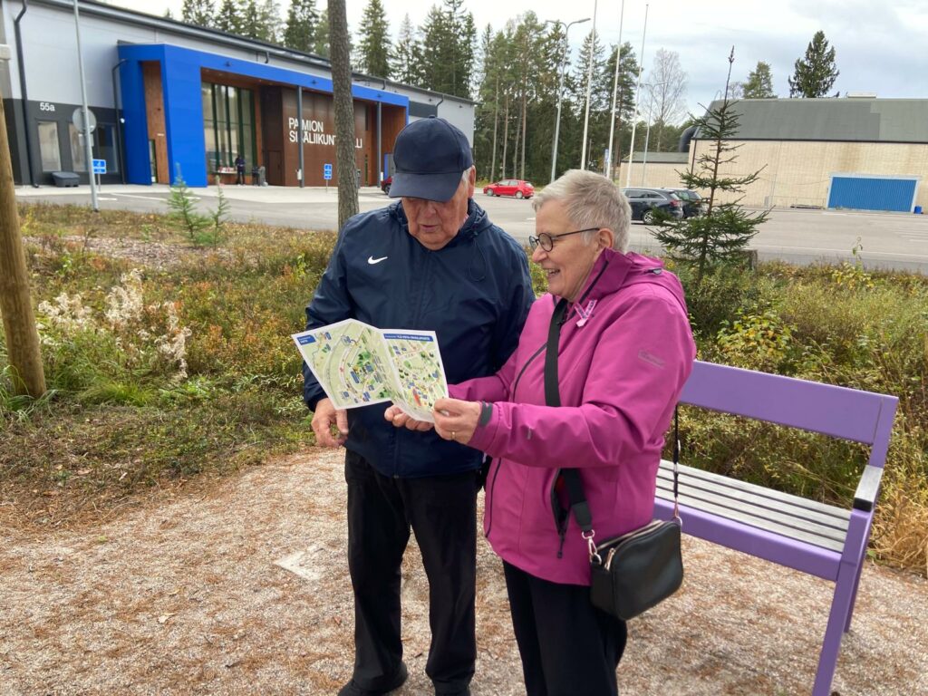 Mies ja nainen seisovat ulkona penkin edessä. Penkin takana näkyy Paimion sisäliikuntahalli sekä sen pysäköintipaikka. Nainen esittelee kädessään olevaa esitettä miehelle.