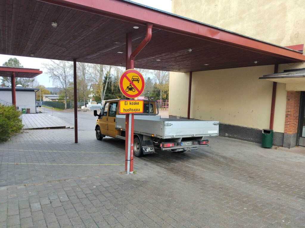 Kahden rakennuksen välillä on katos, jonka alla huoltoauto. Lisäksi kuvan keskellä pyskäköintikieltoa osoittava liikennemerkki.