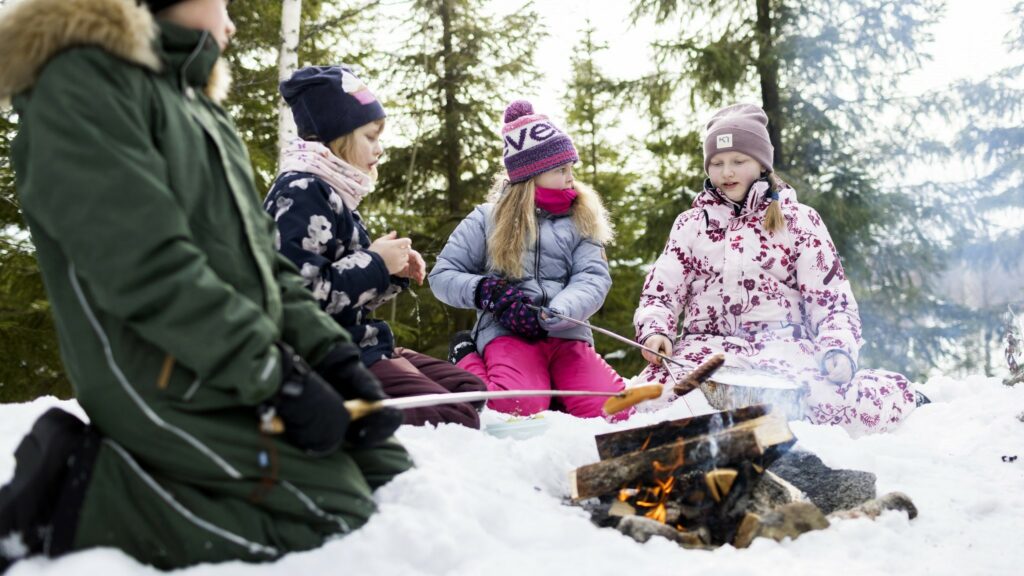 Talvisessa metsässä lapsia nuotion ympärillä paistamassa makkaraa.