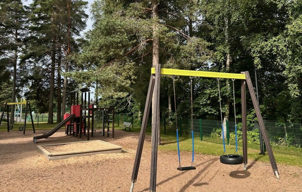 Urheilupuiston leikkipaikan keinut, hiekkalaatikko ja liukumäki kuvattuna kesäaikaan.
